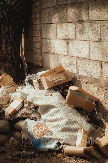 A pile of discarded cardboard boxes, some flattened and others partially intact, along with crumpled paper and plastic packaging materials, is situated on the ground near a concrete brick wall and a tree trunk. The cardboard boxes are in various sizes, with some showing printed labels and barcodes, and they are stacked haphazardly alongside a white, woven sack containing additional rubbish. The surrounding environment appears to be an outdoor area, possibly a back alley or a side yard, with loose gravel and rocks beneath the waste. Visible in the background is a brick wall with a rough texture and a shaded area created by the nearby tree, highlighting a typical scene of waste awaiting collection. Waste Disposal Battersea provides private rubbish removal services, demonstrated here by the accumulation of general waste that could be targeted for efficient collection and disposal, supporting environmentally responsible waste management outside of standard council services.