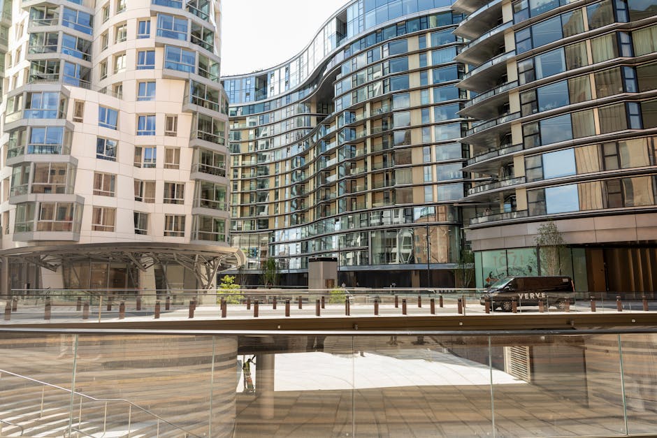 The image displays a modern apartment complex with a distinctive curved glass facade and alternating sections of beige and grey exterior panels. Multiple balconies with glass railings extend from each unit, some with visible patio furniture, and large windows provide ample natural light to the interior. The building's design features smooth, rounded edges and reflective glass surfaces, creating a contemporary aesthetic. In front of the building, there is a wide, paved area with a low metal and glass railing separating an open public space from the street, which has a row of short wooden posts along the edge. A black van is parked on the street to the right, partially obscured by the building, and there are some small trees and shrubs near the entrance, indicating landscaped features. The image is taken during daylight, with clear lighting enhancing the reflective quality of the glass, and the overall scene depicts an urban residential environment. This setting aligns with the context of private waste handling and alternative rubbish collection services commonly used in multi-unit residential buildings, similar to those offered by Waste Disposal Battersea.