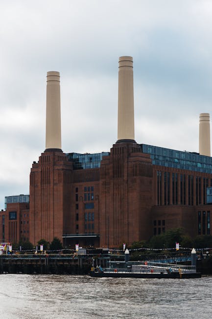 The image features a large, historic power station building with a reddish-brown brick facade, situated along a riverbank. The structure has a prominent, rectangular form with vertical decorative elements and multiple windows arranged in a grid pattern. Four tall, cylindrical chimneys extend upward from the roof, each with a smooth, light-colored surface and capped with a rounded top. The building’s surface shows signs of age, with some areas of weathering, and is partly covered by modern blue-tinted glass extensions at the top, contrasting with the original brickwork. In the foreground, the river’s surface appears slightly rippled, with a narrow boat or barge docked along the quay, indicating a setting that may be associated with industrial or urban waste management activity. Surrounding the water, small trees and pavement are visible, adding some greenery and urban context. Overcast skies above cast diffuse lighting over the scene, emphasizing the industrial character of the building. This setting aligns with the idea of private waste handling or on-site clearance, similar to rubbish removal services provided by companies like Waste Disposal Battersea.
