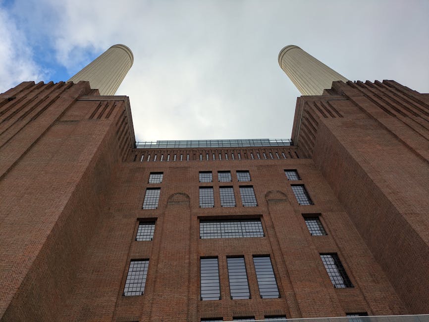 A low-angle view of a large, historic brick building with a symmetrical facade, featuring numerous rectangular windows with black frames. The structure includes prominent vertical brick columns on either side that extend upward and support two tall, cylindrical, cream-colored chimneys or towers positioned to the left and right of the building. The brickwork has a reddish-brown hue with a textured surface, and the windows are set in recessed arches, adding architectural detail. The sky above is partly cloudy with patchy blue visible, casting soft natural light on the scene. The photograph captures the building's imposing height from a perspective that emphasizes its grandeur and the industrial or historical character, typical of structures that might be associated with old factories or civic landmarks, often involved in waste or material processing, relevant to waste disposal or rubbish removal contexts carried out by companies like Waste Disposal Battersea.