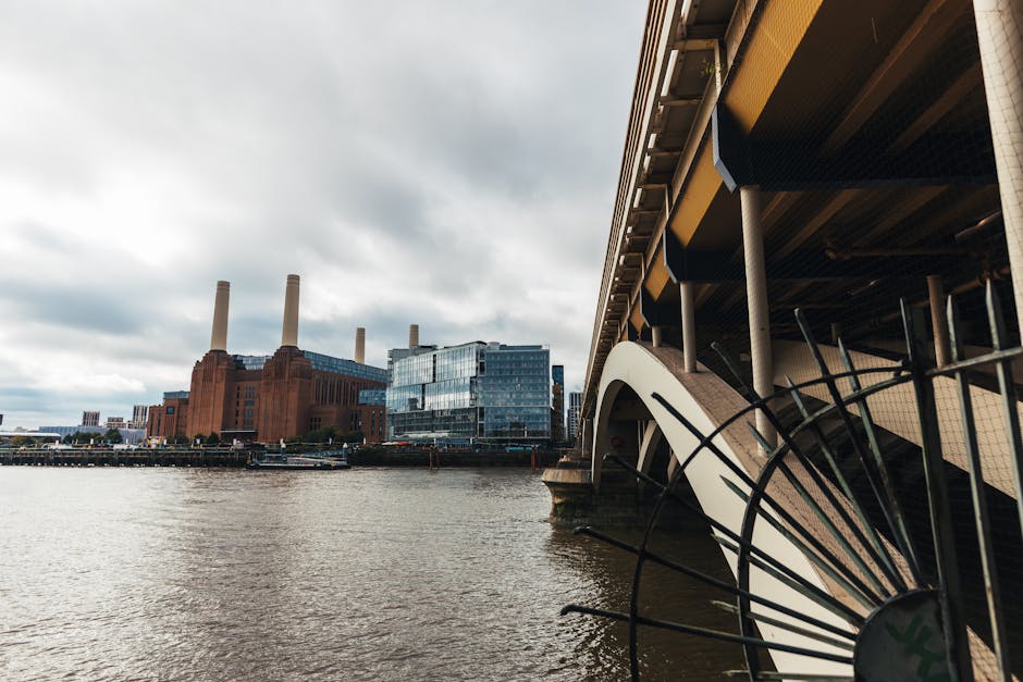The image features a large, historic power station building with a reddish-brown brick facade, situated along a riverbank. The structure has a prominent, rectangular form with vertical decorative elements and multiple windows arranged in a grid pattern. Four tall, cylindrical chimneys extend upward from the roof, each with a smooth, light-colored surface and capped with a rounded top. The building’s surface shows signs of age, with some areas of weathering, and is partly covered by modern blue-tinted glass extensions at the top, contrasting with the original brickwork. In the foreground, the river’s surface appears slightly rippled, with a narrow boat or barge docked along the quay, indicating a setting that may be associated with industrial or urban waste management activity. Surrounding the water, small trees and pavement are visible, adding some greenery and urban context. Overcast skies above cast diffuse lighting over the scene, emphasizing the industrial character of the building. This setting aligns with the idea of private waste handling or on-site clearance, similar to rubbish removal services provided by companies like Waste Disposal Battersea.