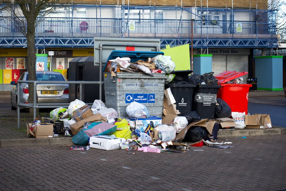 A large collection of overflowing rubbish and waste materials is piled on a paved sidewalk in front of a commercial premises. The waste includes brown cardboard boxes, plastic shopping bags, black refuse sacks, and loose paper and packaging scattered around. A prominent grey recycling bin labeled 'mixed paper & card' is open with crumpled papers and cardboard protruding from inside. Nearby, black and red waste bins are also filled with refuse, some overflowing with debris spilling onto the ground. Behind the waste piles, a metal railing runs parallel to the sidewalk, and a parked silver car is visible on the left side of the image. The background features a storefront with a yellow and red sign, and above it, a building with glass windows and blue metal construction barriers on the upper floor. The scene is set in an urban area, capturing the impact of an uncollected rubbish collection, indicative of private waste handling or alternative rubbish removal services. Waste Disposal Battersea occasionally manages such waste clearing tasks, as depicted here, in the context of non-local authority rubbish collection.