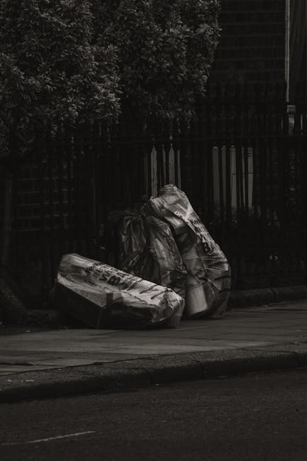 A close-up view of three crumpled and partially torn plastic rubbish bags placed on the sidewalk beside a black metal fence with vertical bars. The bags are filled with waste materials, with some printed text visible on the packaging, and are situated near a curb on a paved street. In the background, greenery from a bush or tree extends above the fence, and a dark building or structure is partially visible behind the foliage. The scene is captured in low lighting or shade, emphasizing the texture and creases of the bags. This image illustrates the type of waste collection that may be arranged through private waste disposal services such as Waste Disposal Battersea, providing an alternative to council-run rubbish collection methods nearby the Battersea area, and emphasizes the importance of proper rubbish handling and disposal at the roadside in urban settings.