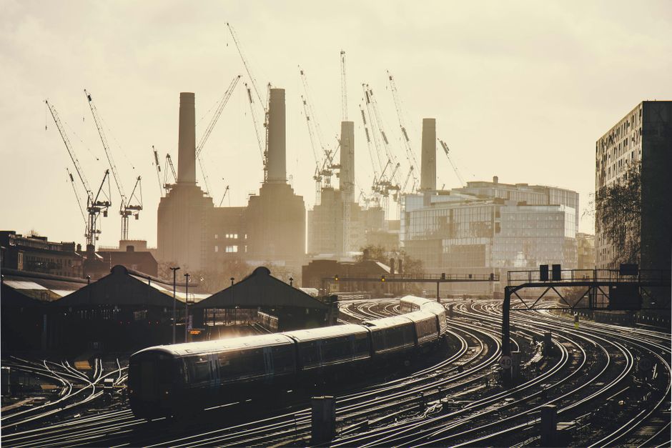 A black modern train with a sleek, rounded design is positioned on curved railway tracks in the foreground, with metallic surfaces reflecting ambient light. The tracks, made of steel rails and wooden sleepers, expand and diverge across the scene, leading towards a dense cluster of industrial and construction sites in the background. Behind the tracks, there are two large, dark-colored canopy structures, possibly for railway platforms or storage areas, with a hint of orange lighting underneath. Further in the distance, multiple tall construction cranes with long, articulated arms and cables are visible, positioned against a hazy sky, indicating ongoing development at a large construction site with partially completed buildings. The backdrop features high-rise buildings with a variety of window arrangements and finishes, and the overall atmosphere is characterized by diffused natural lighting, possibly early morning or late afternoon, creating a subdued, muted tone. This scene exemplifies urban infrastructure and construction activity, indirectly connecting to the themes of bulk waste management or debris clearance handled through independent waste disposal services like Waste Disposal Battersea, servicing the Battersea area with efficient rubbish removal solutions.