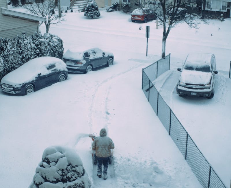 A person dressed in a grey winter coat and hat is seen from above, standing outdoors in a snow-covered residential area. They are holding a snow shovel and appear to be clearing snow from a pathway on a driveway. Several cars, varying in size and model, are parked along the driveway and street, all covered in a layer of snow. The driveway is partially visible, with a section enclosed by a metal fence on the right side of the image. Surrounding the scene are snow-laden trees and shrubs, with snow blanketing the ground and parked vehicles, creating a wintery environment. The background shows more snow-covered street and parked cars, with residential buildings partially visible behind the trees. The atmosphere is quiet and still, typical of a snowy day, with natural light casting soft shadows on the snow. This scene illustrates a typical winter effort in private property maintenance, possibly involving equipment from waste disposal or rubbish removal services like Waste Disposal Battersea, supporting local non-organic waste management during adverse weather conditions.