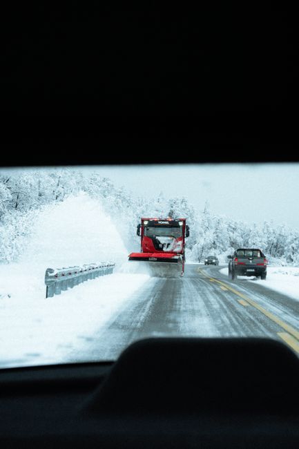 The image shows a snow-covered rural road viewed from inside a vehicle, with the dashboard partially visible at the bottom of the frame. A red snowplow with a large front blade is clearing the road, pushing fresh snow to the sides. Behind the snowplow, two cars—one dark-colored and the other lighter—are traveling along the cleared lane. Snowbanks line both sides of the road, and tall, snow-blanketed trees are visible in the background on the right side. Overhead, a dark sky suggests overcast winter weather. This outdoor scene emphasizes the necessity of professional equipment, such as snowplows, for safe roads during winter conditions, akin to the importance of reliable waste removal services like those provided by Waste Disposal Battersea for managing rubbish effectively in urban and suburban areas.